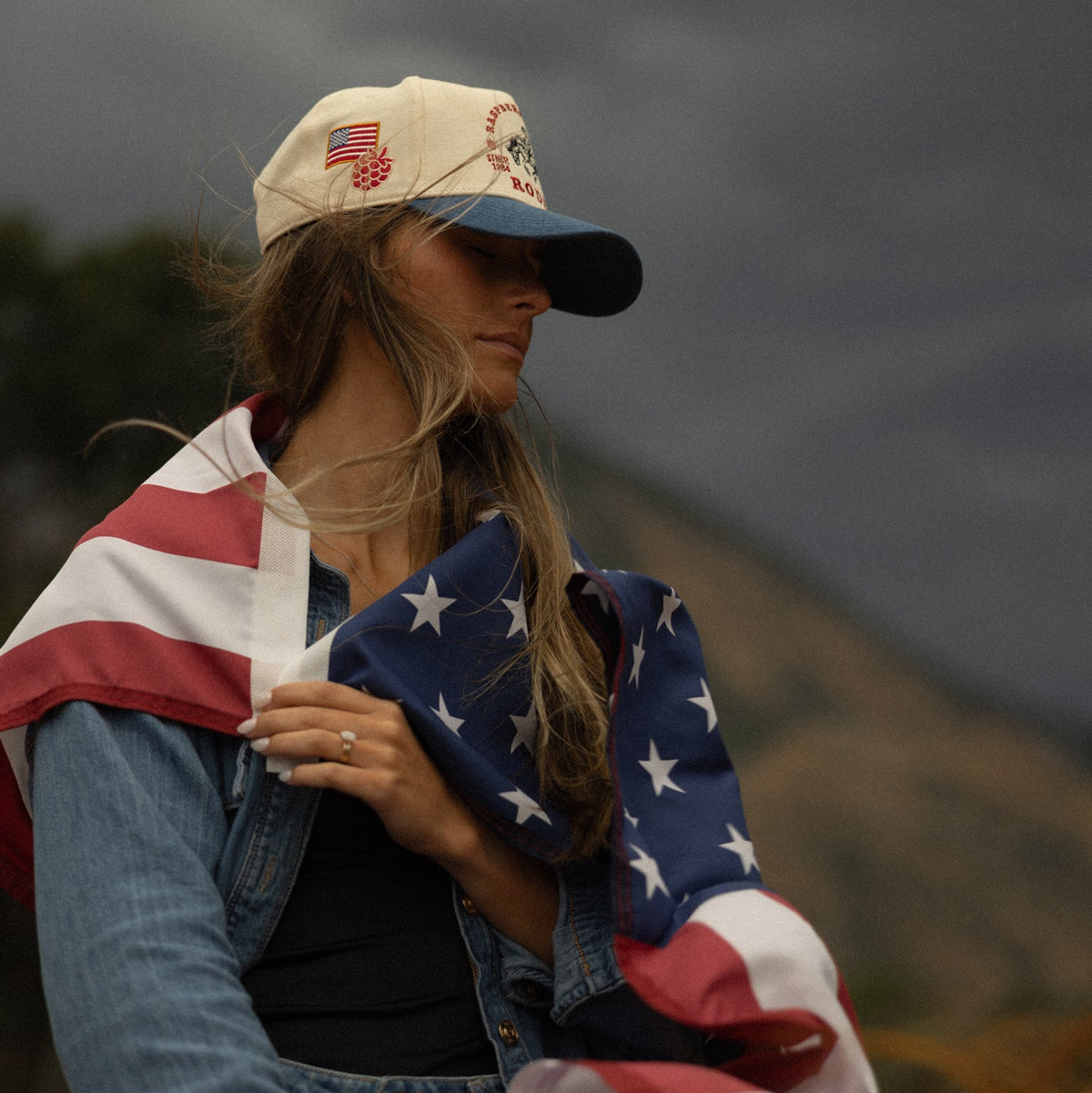 Person wrapped in an American flag with a raspberry days cap, standing outdoors against a dark sky.