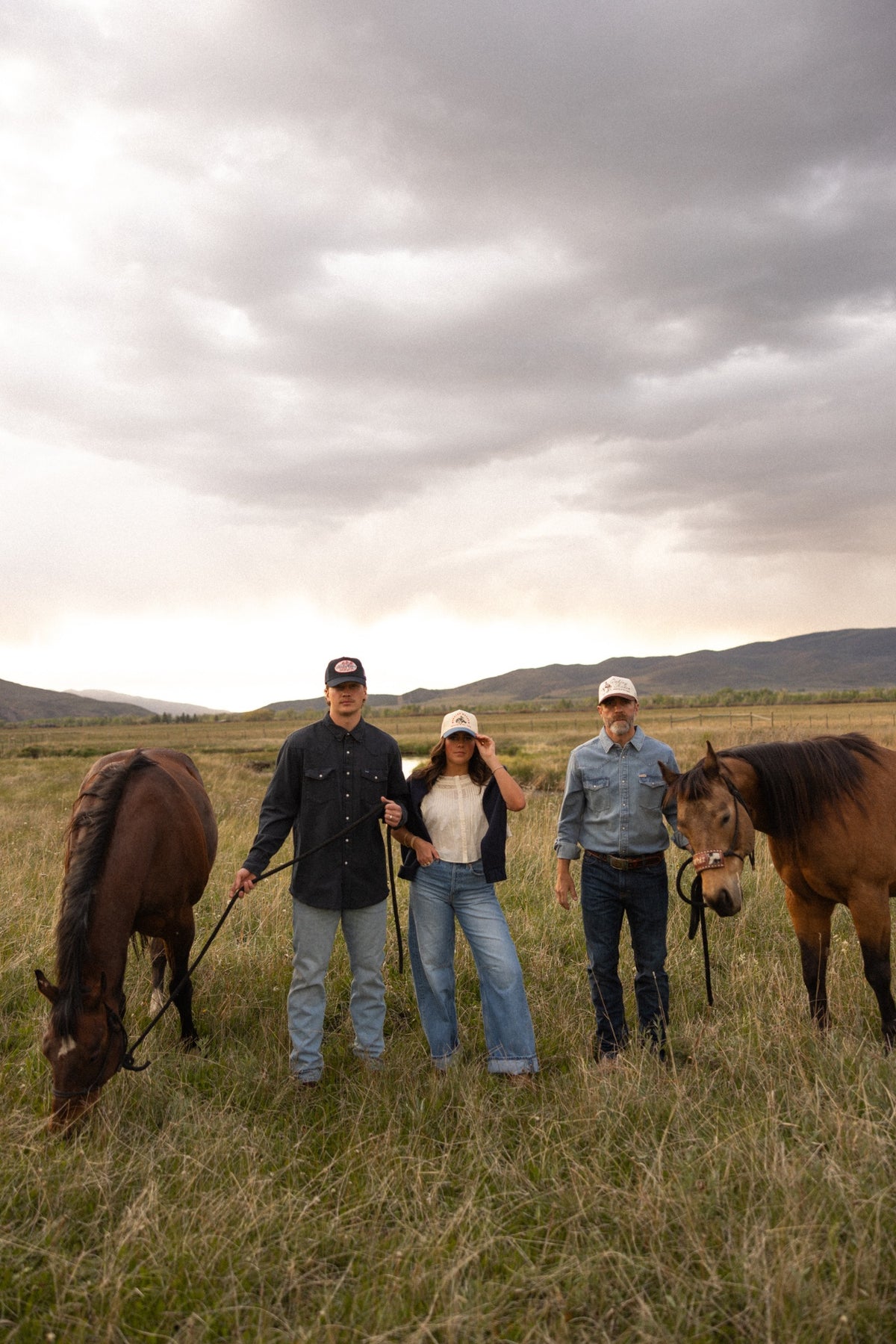 Three models with horses wearing custom Bolt Ranch trucker hats and western shirts under dramatic sky