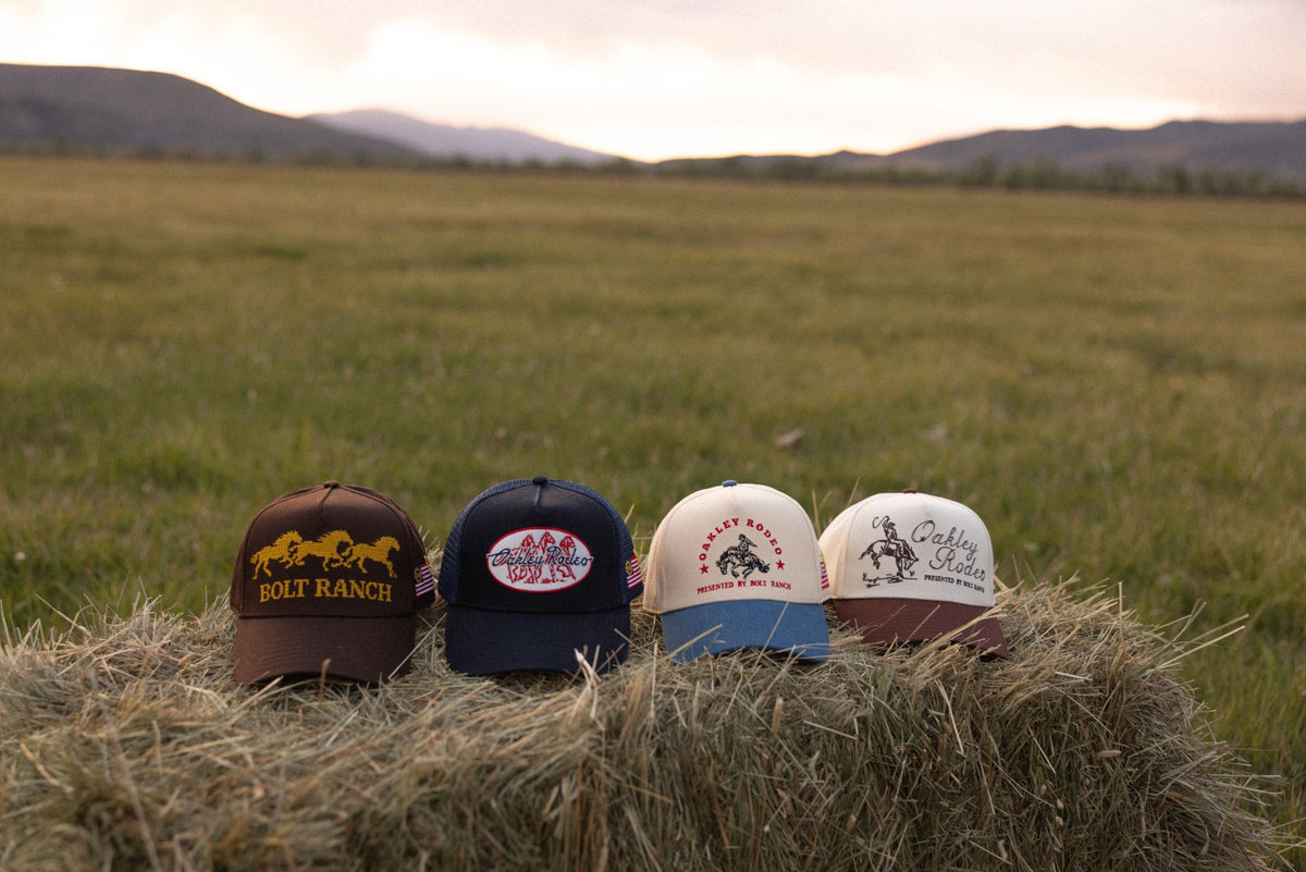 Four Bolt Ranch and Oakley Rodeo hats displayed on a hay bale at sunset