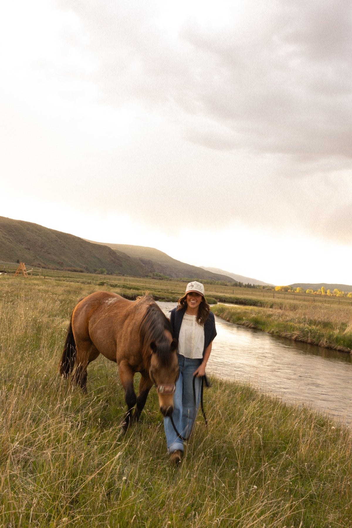 Woman walking a horse along a creek wearing a custom ranch trucker hat at golden hour