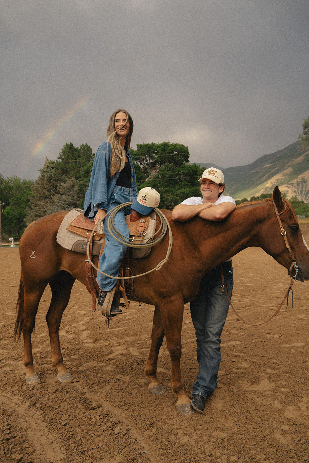 Couple with horse and custom rodeo trucker hats under a rainbow after the storm