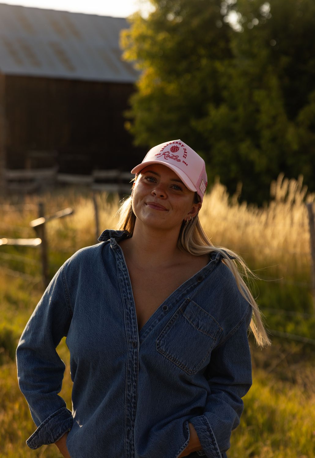 Model in a pink Raspberry Days Rodeo trucker hat and denim in front of a barn