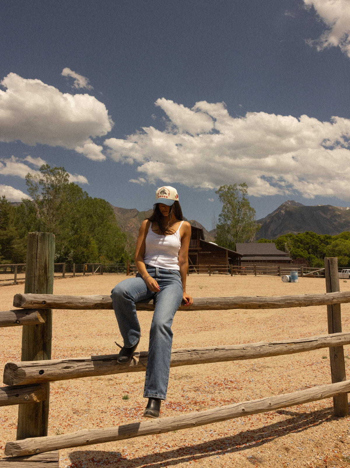 Model on a fence rail in a Strawberry Days Rodeo cap with mountains and barn behind